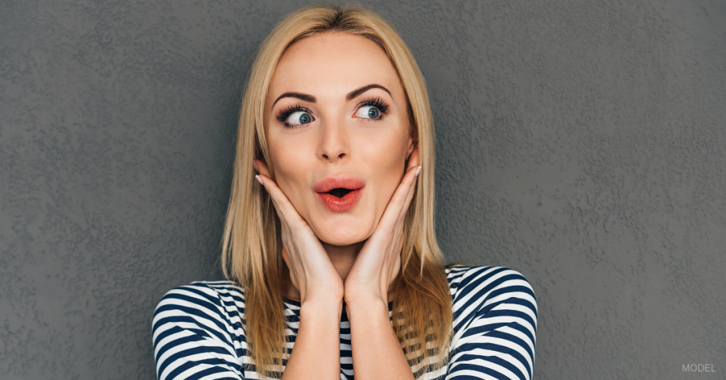 Woman with smooth skin and full, red lips looks surprised with hands on either side of face